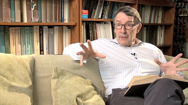 Chris Isham sitting on a sofa with textbook in lap, talking about quantum mechanics