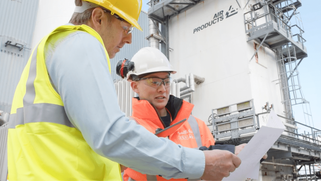 Two workers in hard hats and hi viz vests in front of a tall white Air Products industrial tower