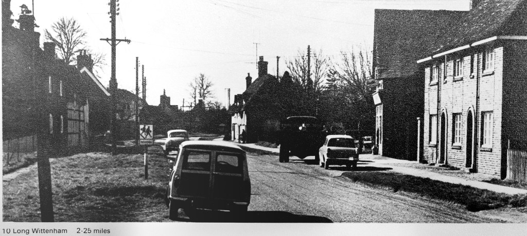 Black and white photo of village street with tall chimney drawn on in distance above houses