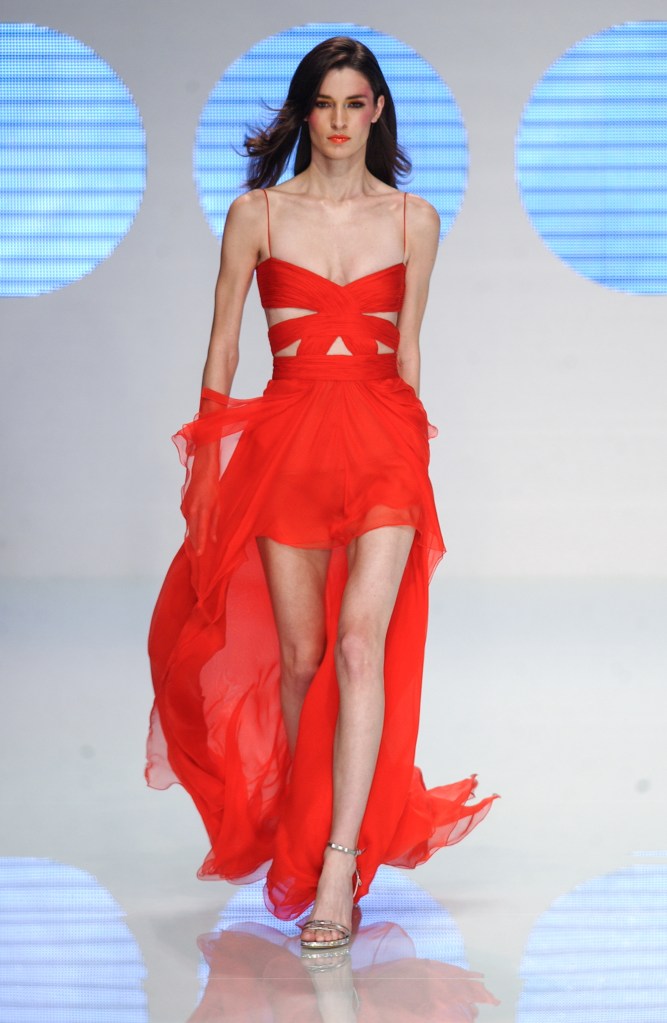 Tall model with black hair wears striking red dress with short skirt and trails behind with wide ribbons crossing across the chest